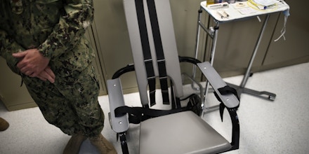FILE - In this photo Nov. 20, 2013 file photo reviewed by the U.S. military, a U.S. Navy nurse stands next to a chair with restraints, used for force-feeding, and a tray displaying nutritional shakes, a tube for feeding through the nose, and lubricants, including a jar of olive oil, during a tour of the detainee hospital at Guantanamo Bay Naval Base in Cuba. Attorneys for a Syrian prisoner have begun studying hours of video showing him being removed from his cell, placed in a restraint chair and fed by a tube with liquid nutrients. They are looking for evidence of what he has portrayed to them as abusive force-feeding, akin to torture, during the months that he has participated in a hunger strike that drew the attention of President Barack Obama and led to a renewed effort to close the prison. (AP Photo/Charles Dharapak, File)