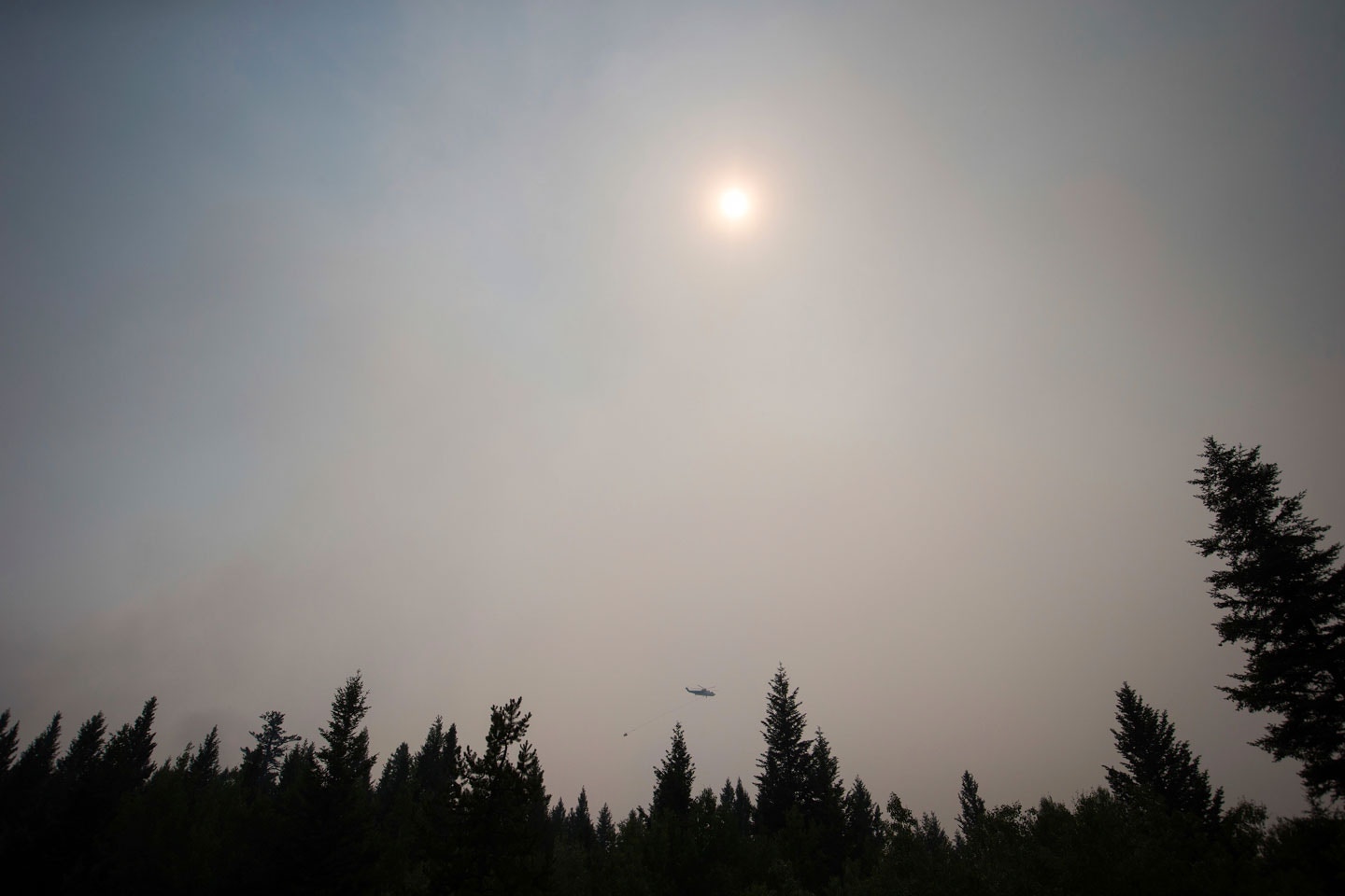Smoke obscures the sun as a helicopter carrying a bucket battles the Gustafsen wildfire near 100 Mile House, B.C., on Saturday July 8, 2017. More than 180 fires were burning, many considered out of control, as the B.C. government declared a provincewide state of emergency to co-ordinate the crisis response. Officials said buildings have been destroyed, but they did not release numbers. The BC Wildfire Service says over 173 fires were reported on Friday alone as lightning storms rolled over several parts of B.C. (Darryl Dyck/The Canadian Press via AP)