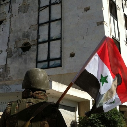 A Syrian army's soldier a national flag featuring Syria's President Bachar al-Assad in front of a building left in ruins with on June 5, 2013 in the city of Qusayr in Syria's central Homs province, after the Syrian government forces seized total control of the city and the surrounding region. The Syrian army ousted rebels from the strategic town of Qusayr after a blistering 17-day assault led by Hezbollah fighters, scoring a major battlefield success in a war that has killed at least 94,000 people.   AFP PHOTO / STR        (Photo credit should read -/AFP/Getty Images)