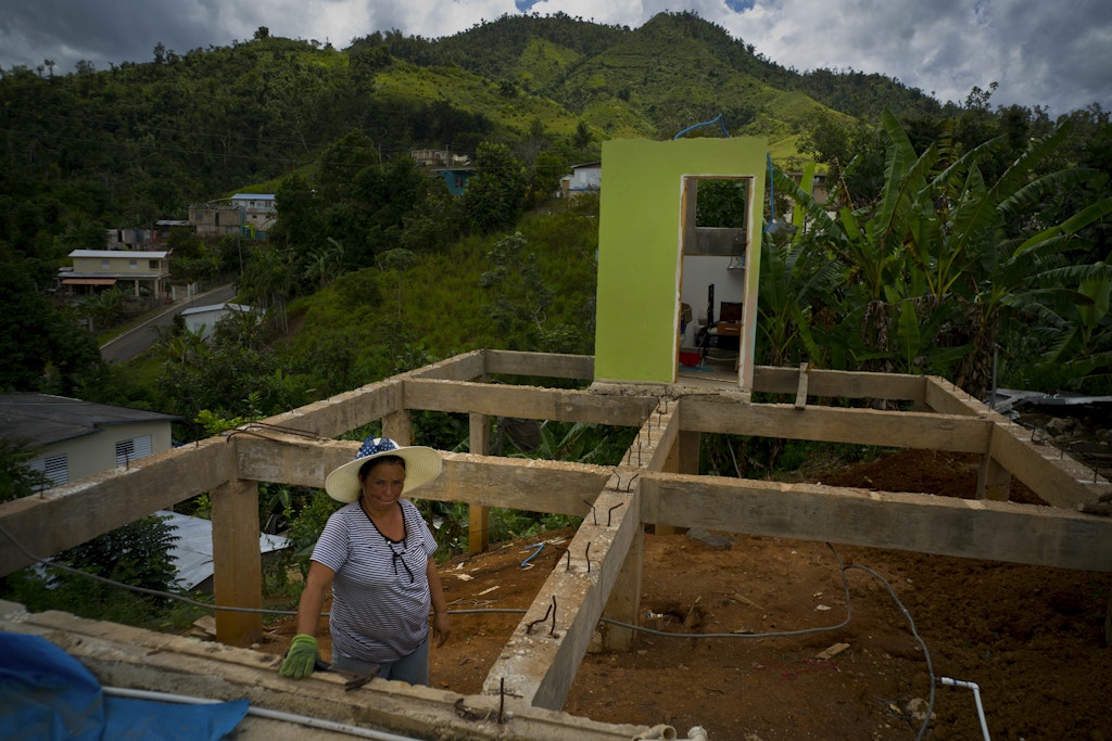 In this Sept. 8, 2018 photo, Alma Morales Rosario poses for a portrait between the beams of her home being rebuilt after it was destroyed by Hurricane Maria one year ago in the San Lorenzo neighborhood of Morovis, Puerto Rico. Rosario, who is incapacitated by diabetes and a blood disease, took a loan to upgrade her home before the storm hit, and lost everything. After the storm, Rosario rented a home until she could no longer afford it on her monthly $598 dollar pension and now splits her time living with her mother and daughter. Rosario said she already spent her $7,000 dollars of FEMA aid, and is now using money from a relative, who is also helping her with the labor of rebuilding her home, but says she knows there's not enough money for all the materials. "I hope with God's help to have the house closed on the outside, walls and ceiling in November. But if it's not possible, I'll make a room with the wood I have under the structure and live there until I can finish it. I never thought this was going to happen to me," she said. (AP Photo/Ramon Espinosa)