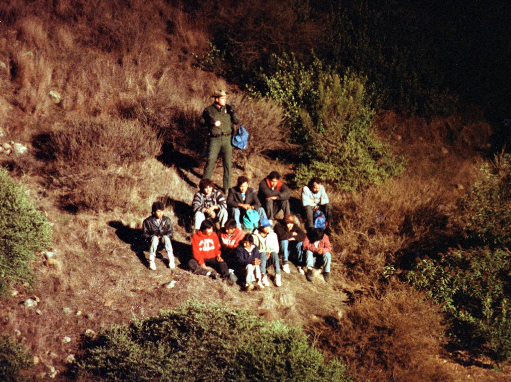 Imigrantes mexicanos são revistados pela Patrulha de Fronteira dos EUA numa íngreme encosta de cânion, iluminada por um helicóptero da Patrulha de Fronteira.