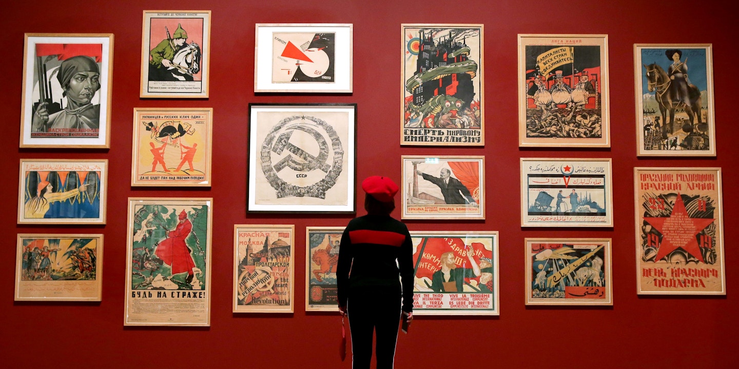 A visitor poses in front of a mural with various posters including: artist Adolf Strakhov's 'Emancipated Woman: Build socialism!' c. 1926 (top-L) and artist Valentin Shcherbakov's 'A Spectre is Haunting Europe, the Spectre of Communism' c. 1924, during the 'Red Star over Russia: A revolution in Visual Culture 1905-55' exhibition at the Tate Modern in London on November 7, 2017. / AFP PHOTO / Daniel LEAL-OLIVAS / RESTRICTED TO EDITORIAL USE - MANDATORY MENTION OF THE ARTIST UPON PUBLICATION - TO ILLUSTRATE THE EVENT AS SPECIFIED IN THE CAPTION (Photo credit should read DANIEL LEAL-OLIVAS/AFP via Getty Images)