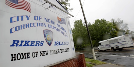 FILE - In this May 17, 2011  file photo, a New York City Department of Corrections bus passes the sign near the gate at the Rikers Island jail complex in New York.  Four correctional officers at Rikers Island jail have been suspended without pay after they allegedly stood by and failed to stop a teenage inmate from hanging himself on Thanksgiving, leaving him hospitalized in intensive care. Nicholas Feliciano, 18, remained unconscious in a hospital prison ward Wednesday, Dec. 4, 2019 a week after he nearly died in his Rikers Island jail cell, according to the Legal Aid Society, the public defender organization that represents him in court.  (AP Photo/Seth Wenig, File)