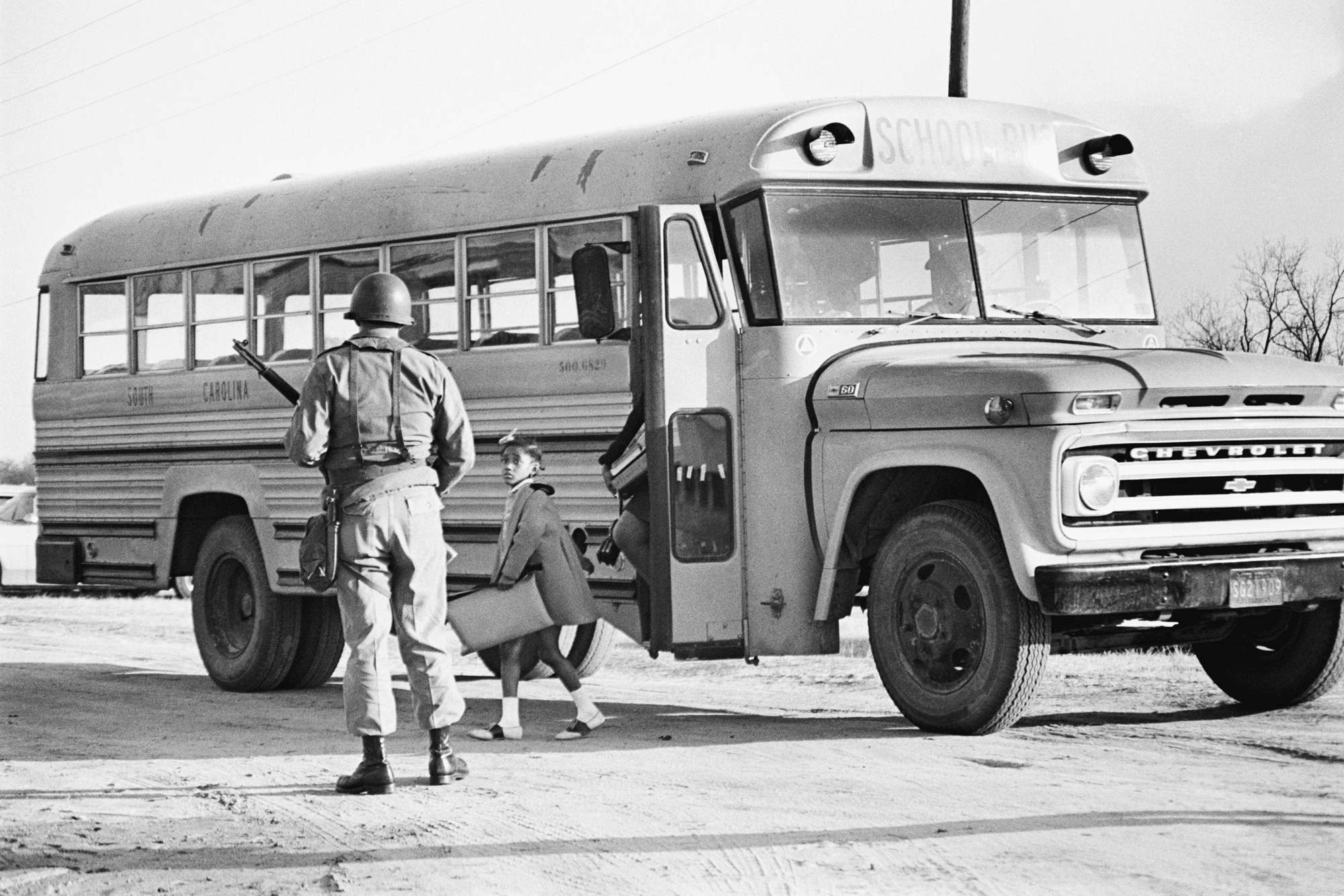 A South Carolina National Guardsman meets a school bus as it arrives with African American students at the Lamar School. A little girl watches the guardsman with uncertainty. A mob of whites from the town of Lamar resisted school integration by turning over buses as they arrived at the school. | Location: Lamar School, Lamar, South Carolina, USA.