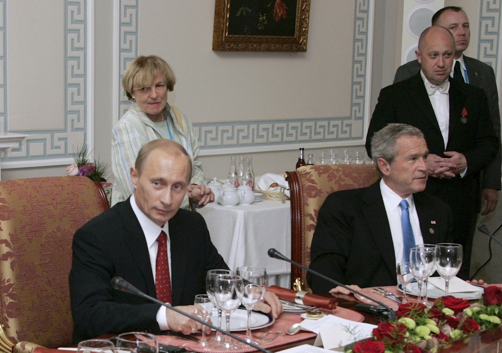FILE - In this Monday, July 19, 2006, file photo, Russian President Vladimir Putin, left, and U.S. President George W. Bush attend a working dinner with the other leaders of the G8 nations, while Russian businessman Yevgeny Prigozhin stands, right, in St. Petersburg , Russia.  While Russian officials have downplayed a US indictment charging 13 Russians with meddling in the 2016 US presidential election through an elaborate social media campaign, former Internet trolls employed at the same facility see them as well-founded.  The indictment claimed that Yevgeny Prigozhin _ a wealthy entrepreneur and restaurateur called "Putin's chef" _ funneled money to set up the troll factory that sent operatives to the US, created fictitious social media accounts and used them to spread tendentious messages.  (Sergei Zhukov, Sputnik, Kremlin Pool Photo via AP, file)